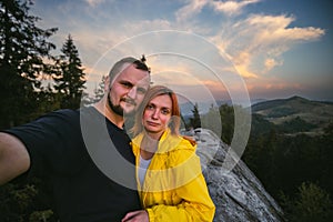 Couple hikers taking a selfie on top of a mountain, summer sunset time. Man and woman looking at beautiful inspirational landscape