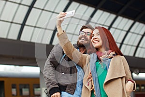 Couple of hipster travelers photographing a selfie with a smartphone in a train station. Travel concept. Mobile