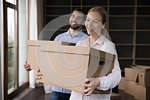 Couple holding boxes with belongings posing in unfurnished living room