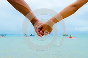 Couple holding hands having the sea as a background. View of the arms in the center. Some boats in blur.