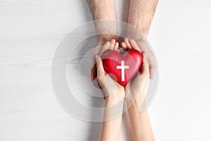 Couple holding heart with cross symbol on background, top view. Christian religion
