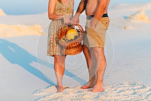 Couple holding a wicker basket with fruit, man and woman having a picnic on the white sand beach or in the desert or in the sand d