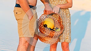 Couple holding a wicker basket with fruit, man and woman having a picnic on the white sand beach or in the desert or in the sand d