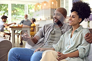 Couple At Home On Sofa Streaming Show Or Movie To TV With Multi-Generation Family In Background