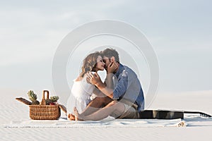 Couple hugging while sitting on blanket with basket of fruits and acoustic guitar on beach
