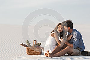 Couple hugging while sitting on blanket with basket of fruits and acoustic guitar on beach