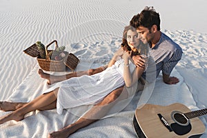 Couple hugging while sitting on blanket with basket of fruits and acoustic guitar on beach