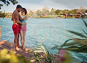 Couple hugging. Young man and woman in embrace, standing by water on wooden jetty. Blue water and houses in background.