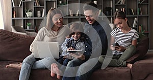 Couple and little children relax on couch using diverse devices
