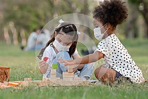 Couple little girls African and Caucasian kids wear face mask while sitting and playing wooden blocks toy in green park together