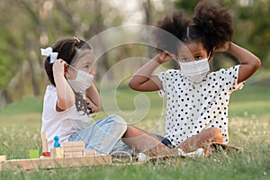 Couple little girls African and Caucasian kids wearing face mask while sitting and playing wooden blocks in green park together