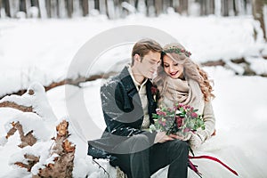 Couple in love with a bouquet are sitting on the log on background of the snowy forest. Winter wedding. Artwork