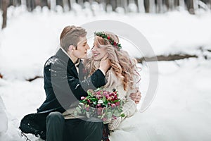 Couple in love with a bouquet are sitting on the log on background of the snowy forest. Winter wedding. Artwork