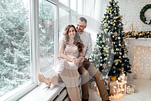 Couple in love in festive clothers sitting and hugging near the large window and Christmas tree in decorated studio. happy holiday