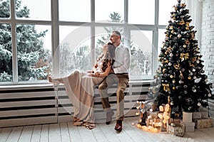 Couple in love in festive clothers sitting and hugging near the large window and Christmas tree in decorated studio. happy holiday