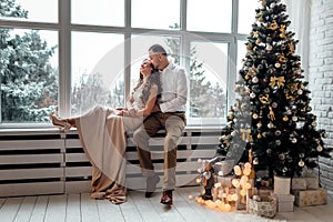 Couple in love in festive clothers sitting and hugging near the large window and Christmas tree in decorated studio. happy holiday photo