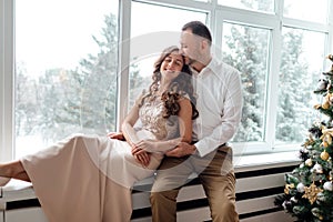 Couple in love in festive clothers sitting and hugging near the large window and Christmas tree in decorated studio. happy holiday