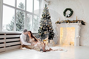 Couple in love in festive clothes sitting and hugging near the large window and Christmas tree in decorated studio. happy holiday
