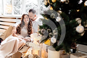 Couple in love in festive clothes sitting and hugging near the large window and Christmas tree in decorated studio. happy holiday