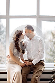 Couple in love in festive clothes sitting and hugging near the large window and Christmas tree in decorated studio. happy holiday