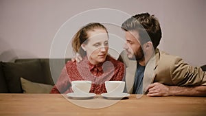 A couple in love having coffee, sitting on a sofa, talking to each other and happily smiling.