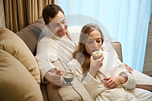 Couple in love having lunch sitting in bathrobes on sofa