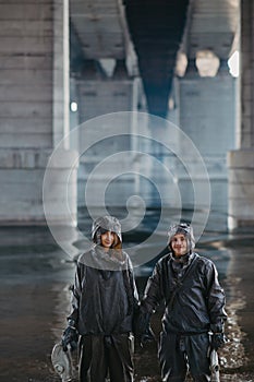Couple in love holds hands and stands under bridge in NBC protective suits and gas masks in their hands