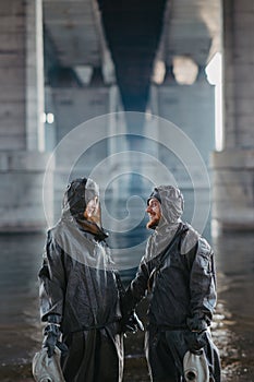 Couple in love holds hands and stands under bridge in NBC protective suits and gas masks in their hands