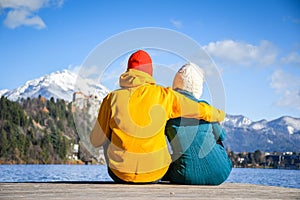 Couple in love hugging together with colorful cloths sitting and relaxing on a wooden pier on a clear sky sunny winter day view fr