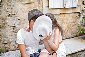 Couple in love kissing behind white female hat on the stone bench in old city. Young man and woman hugging. Summer sunny day