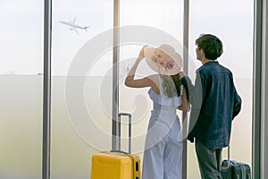Couple love is looking at flying plain in sky. They are standing near window at airport and holding hands