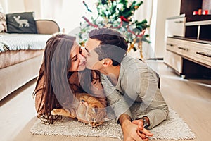 Couple in love lying by Christmas tree and playing with cat at home. Man and woman kissing