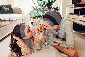 Couple in love lying by Christmas tree and playing with cat at home. Man and woman kissing pet