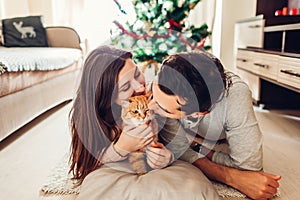 Couple in love lying by Christmas tree and playing with cat at home. Man and woman relaxing