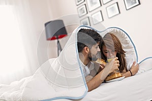 Couple in love lying under the sheets, having fresh croissants and orange juice for breakfast in bed and enjoying the weekend