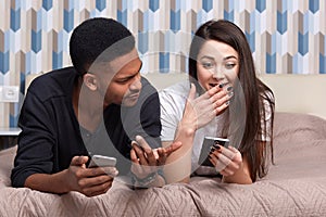 Couple in love relaxing together while lieing on bed. Young man and woman looking at mobile phone with astinished facial
