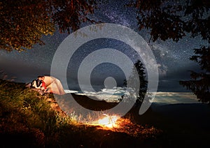 Couple in love sits on hill near tent enjoying burning fire under night sky strewn with bright stars