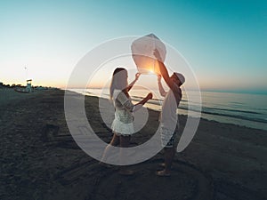 Couple of lovers lighting sky lantern on the beach at sunset - Young people celebrating their relation anniversary in summer