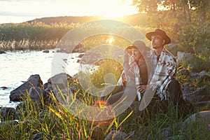 Couple man and woman in felt hats wrapped in whool blanket sitting near lake on sunset. Weekend picnic on nature, local travel,