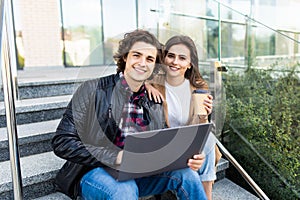 Couple man and woman with paper cup smiling and using laptop while sitting on stairs outdoors
