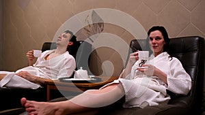 Couple man woman in white bathrobes drinking tea in sauna lying on armchairs.