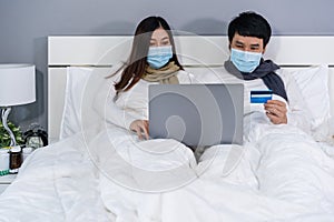 Couple with medical masks using laptop to shopping online with credit card on bed during quarantine pandemic coronaviruscovid-19