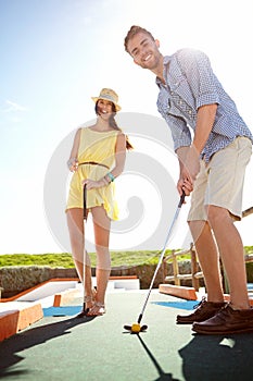 Couple, mini golf and happy man with club, smile and hit ball on date, fun activity and freedom in sunshine. Excited