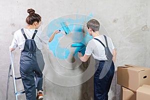 Couple in new home during repair works painting wall together. Happy family holding paint roller painting wall with blue