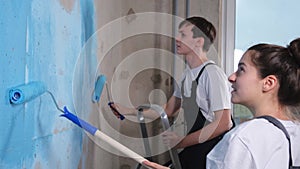 Couple in new home during repair works painting wall together. Happy family holding paint roller painting wall with blue