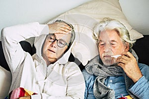 Couple of old aged senior people at home with seasonal winter cold illness disease  sit down on the sof together forever - health