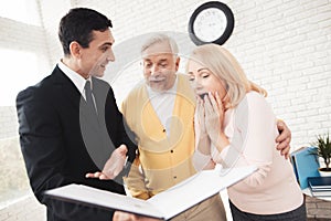 A couple of old people at a reception with a realtor. They stand next to a realtor and shockingly read the form.