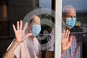 Couple of older people wearing facemasks inside a house looking sadly outside