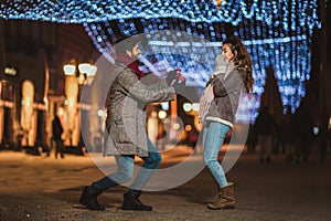 Couple outdoor with holiday`s brights in background. Man presenting gift to woman