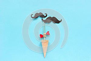 Couple paper mustache props on stick fastened clothespin heart in ice cream waffle cone on blue background. Concept Homosexuality